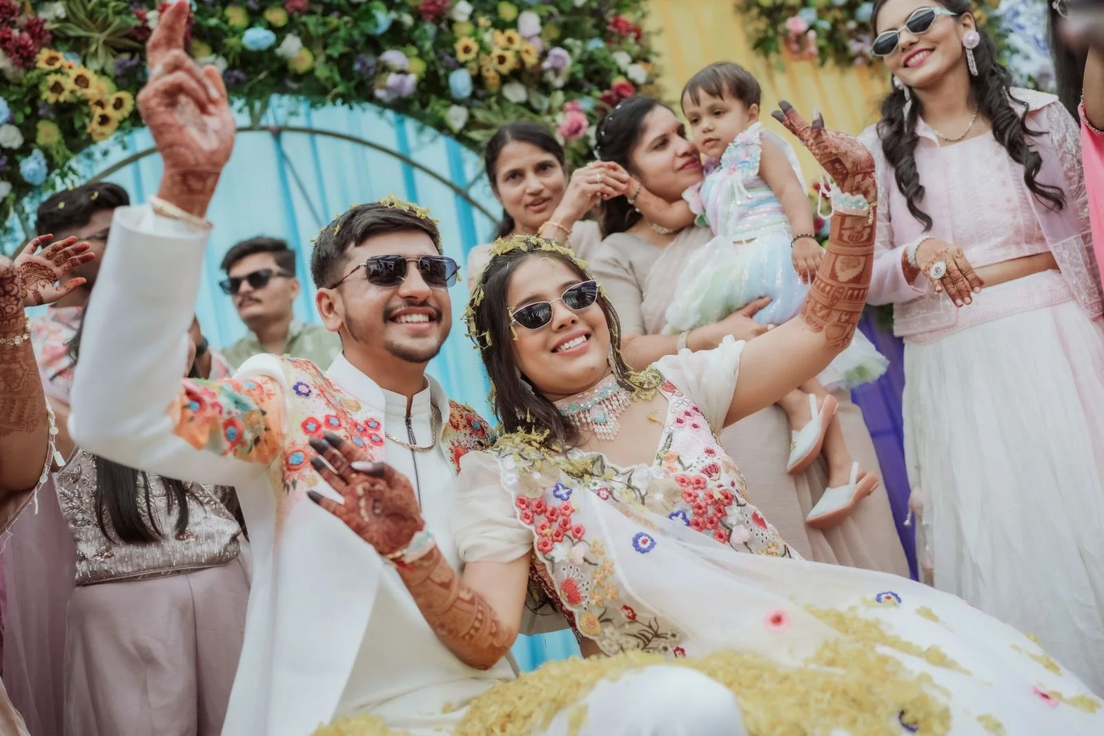 A modern Indian wedding couple and guests celebrating beneath a floral arch.